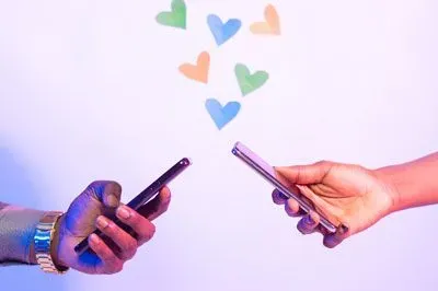 two hands, each holding a cell phone, with artistic hearts floating up between them