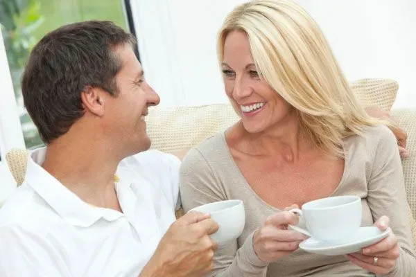 Two 40+ singles enjoying a coffee date and smiling at each other, holding coffee cups and sitting on a couch